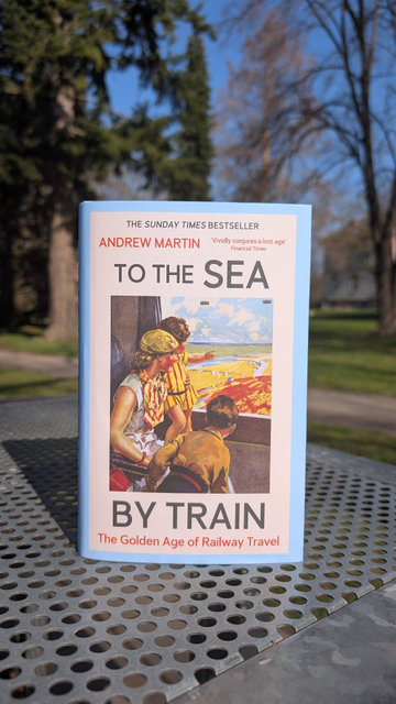 The book "To the Sea by Train" by Andrew Martin, on a metal table in front of trees, green meadow and blue sky