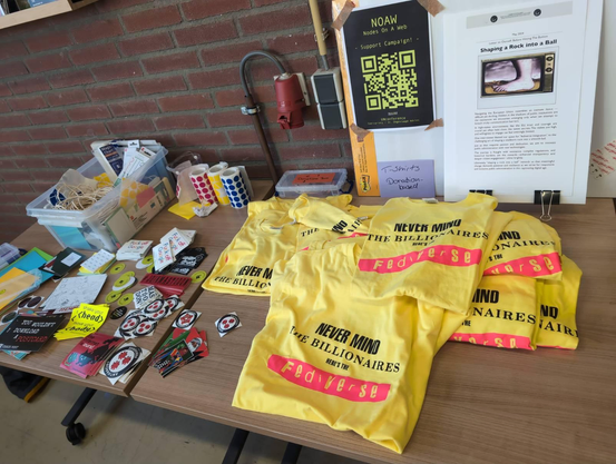 A table with office material, stickers and yellow T-shirts in the style of the sex pistols (black letterson top and a pink invert on the bottom) spelling "Never mind the billionares here's the fediverse". 
Next to them is a purple sign saying "T-Shirts donation-based" and up from it is a A4 QR code asking for support for donation for the NOAW unconference. 
On the right there is story card with a Monty Python reference picture of a foot and the title "Shapping a rock into a ball"
