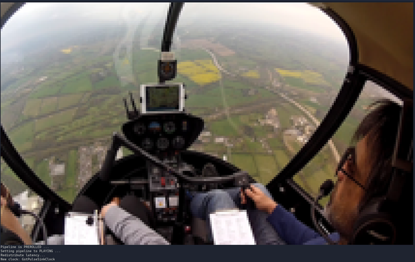 Screenshot of a terminal displaying video output from a GStreamer pipeline rendered directly in the terminal. The frame shows a view from inside a small helicopter cockpit in flight, with the instrument panel in the center. The pilot, seated on the right, is wearing a headset and holding a clipboard. Through the curved windshield, a hazy aerial landscape of fields, roads, and buildings is visible below. Pipeline debug messages appear as text at the bottom of the terminal.