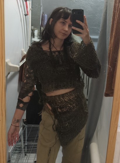 A white person with long dark hair taking a selfie in full length mirror in a bathroom with poorly installed plumbing hoses on the walls. Casually modeling something they knit which is an asymmetrical top with a detached matching piece tied around the waist. Dark green and netlike with another matching, distressed long sleeve shirt underneath.