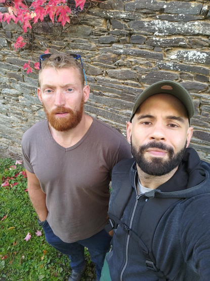 Tristan, le rouquin avec les lunettes de soleil relevées sur la tête & Loïc le brun avec la casquette verte. Tous les deux on met la pose pour ce selfie devant un mur en pierres foncées.