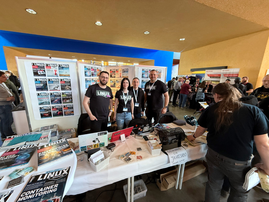 Vier Personen stehen hinter dem Linux-Magazin-Stand bei den Chemnitzer Linux-Tagen und lächeln in die Kamera. Alle tragen schwarze Linux-Magazin-T-Shirts. Auf dem Tisch liegen aktuelle Ausgaben des Linux-Magazins (Titelthema „Container Monitoring"), ein aufgeklapptes Laptop mit NerdZoom-Media-Aufkleber, Sticker, ein kleiner Lautsprecher und ein handgeschriebenes Schild „Gewinne ein Jahresabo". Hinter ihnen zeigen Stellwände Coverabbildungen vergangener Ausgaben. Im Hintergrund ist unter anderem ein Hetzner-Werbebanner zu erkennen.