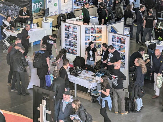Blick von oben auf den Stand von Linux-Magazin  bei den Chemnitzer Linux-Tagen. Drei Stellwände zeigen aktuelle und ältere Titelseiten des Magazins. Mehrere Standmitarbeiter in schwarzen Linux-Magazin-T-Shirts unterhalten sich mit Besucherinnen und Besuchern. Auf dem Tisch liegen Ausgaben der Magazine, ein Schild „Gewinne ein Jahresabo" sowie diverse Sticker und Give-aways. Im Hintergrund sind weitere Messestände zu sehen.