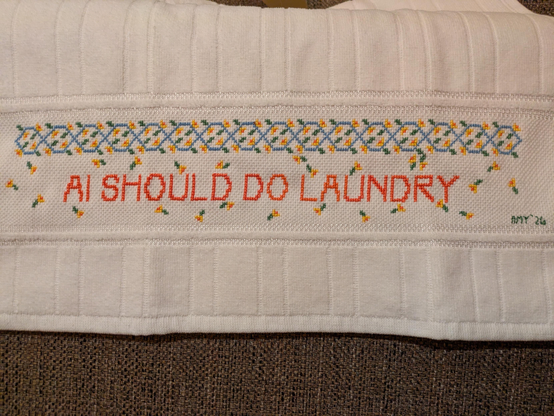An embroidered dish towel that reads "AI SHOULD DO LAUNDRY." There's a floral trellis border and scattered flowers.