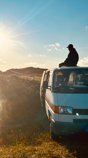 Ein weißer VW-Bus steht in einer hügeligen Graslandschaft (Dünen) im warmen Gegenlicht der tiefstehenden Sonne. Auf dem Dach des Vans sitzt eine Person in Silhouette und schaut entspannt in die Ferne. Die Sonnenstrahlen erzeugen einen hellen Lichtkranz und eine friedliche, abenteuerlustige Atmosphäre.