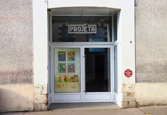 Sur cette photographie, on voit l’entrée de l’atelier Projéta à Nantes. Au-dessus de la porte vitrée, le nom « PROJETA » est visible en lettres blanches sur la vitre supérieure. Dans la vitrine située à gauche de la porte, plusieurs productions, des illustrations et un leporello, sont exposés. Quatre dessins originaux sont accrochés en grille au mur posé à proximité de la vitre, et en dessous se trouvent le leporello posé sur un bloc de bois clair et deux petites images imprimées en sérigraphie.