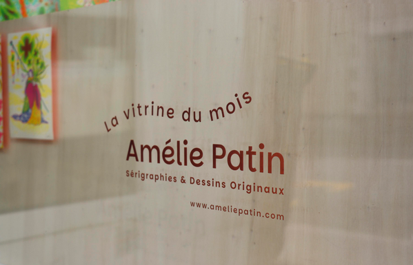 Cette photographie montre un détail de la vitrine. Sur la vitre devant un bloc de bois clair, un texte est imprimé en lettres rouges. On peut lire : « La vitrine du mois / Amélie Patin / Sérigraphies & Dessins Originaux ». En dessous figure l’adresse du site : www.ameliepatin.com. Sur la gauche du texte, derrière la vitre, on peut voir les deux petites illustrations imprimées en sérigraphie.