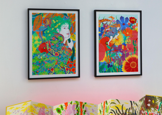 Cette dernière photographie présente deux des dessins aux crayons de couleur encadrés et accrochés au mur, ceux juste au dessus du leporello. Sur celui de gauche, une femme aux cheveux verts est dessinée au milieu d’une végétation dense composée de feuilles et de fleurs. Une petite grenouille verte est visible sur le dos de sa robe. Sur le dessin de droite, on voit une femme sauter parmi de grandes fleurs rouges, jaunes et bleues. En bas de l’image, le haut du leporello déplié est visible.