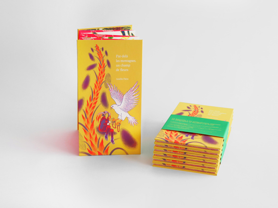 Photographie d’un leporello intitulé Par-delà les montagnes, un champ de fleurs, présenté debout, à côté d’une pile de plusieurs exemplaires fermés et maintenus par un bandeau vert. La couverture jaune montre une scène illustrée avec un oiseau blanc aux ailes déployées, tendant une graine à une personne assise au milieu de grandes plantes. Le leporello a été imprimé en sérigraphie.