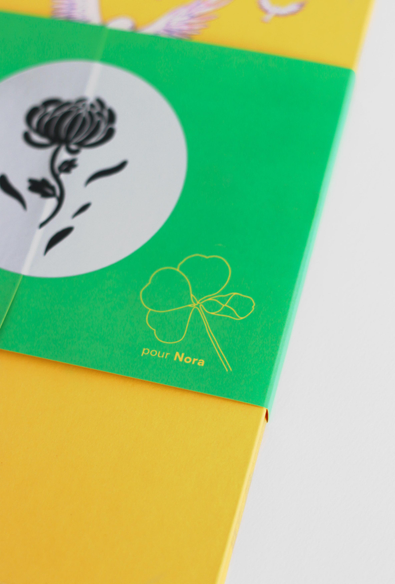 Sur cette photo est présenté le leporello fermé, avec le bandeau vert placé autour. Il est vu de dos, et on peut voir sur le bandeau vert un trèfle à quatre feuille jaune dessiné en ligne claire, accompagné de l’inscription «pour Nora». En haut, un autocollant argenté présente une fleur noire, semblables à celles du motif au verso du leporello. Le fond jaune de la 4e de couverture du leporello est visible.