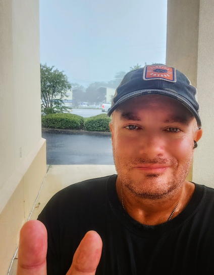 A drenched man standing under a building's overhang during a downpour. Making a peace sign with two fingers. 
