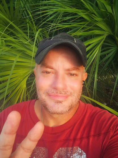 A man standing in front of green palms and making a peace sign with two fingers. 