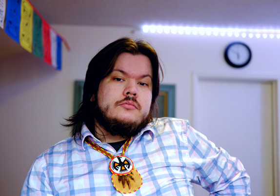 A person with long dark hair and a beard stands with one hand on their hip, looking directly into the camera with a composed, steady expression. They wear a light blue plaid button-up shirt and a beaded thunderbird medallion necklace. A room is visible in the background, with colourful Buddhist prayer flags and a wall clock.