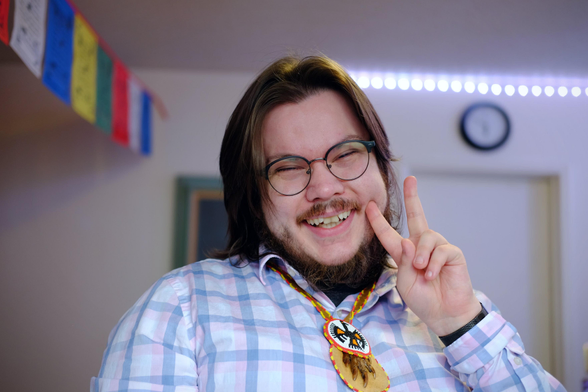 A person with long dark hair, glasses, and a beard grins broadly while holding up a peace sign. They wear a light blue plaid button-up shirt and a beaded medallion necklace featuring a thunderbird design with feather and claw pendants. Colourful Buddhist prayer flags hang in the background.