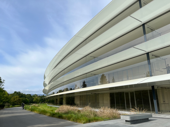 Exterior of Apple Park ring building. A curved glass building under a cloudy sky with white canopy on each level.