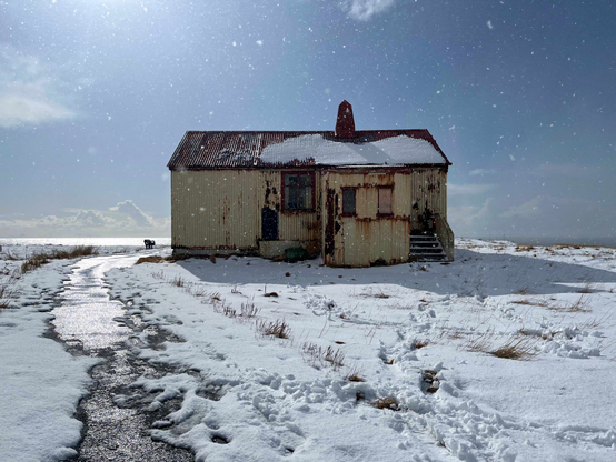A rusty abandoned house surrounded by snow with the sea in the background.