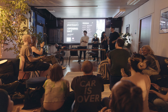 Opening day at pixel.forest() — the team presenting the program to a full room, with the event schedule projected behind them. Warm lighting, plants, and people sitting together on couches and the floor.