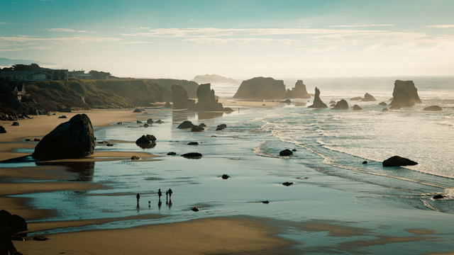A landscape view of a hazy but sunny winter day on the beach with sea stacks all around. A family with a dog can be seen walking in the distance.