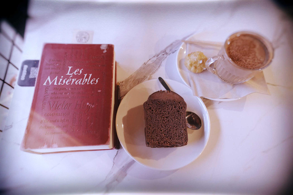 From left to right: a hardcover edition of the novel Les Miserables by Victor Hugo, a slice of chocolate cake on a saucer, and a mug of hot chocolate with a small pastry on a saucer