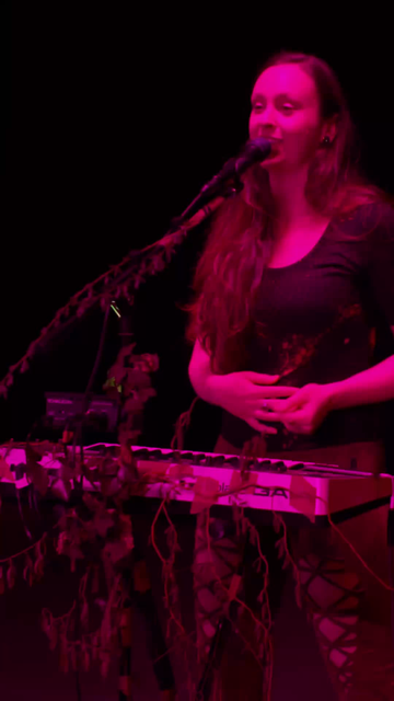 Woman singing on stage with pink stage lights and standing behind her synthesizer which is decorated with plants