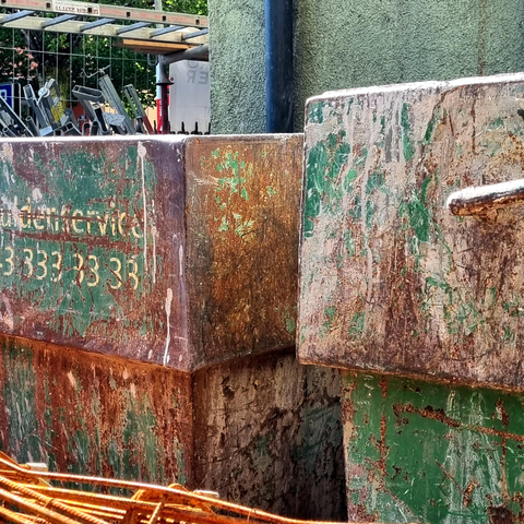 The square image is rendered in shades of rust brown and olive green. It shows a section of a construction site with weathered materials: two shipping containers whose green paint has partially peeled away, revealing patches of white and rust brown; behind them, a green house wall; to the left of that, a ladder lying horizontally; and at the very bottom of the image, rusted metal struts. On one of the containers, “.33333333” is visible in white.