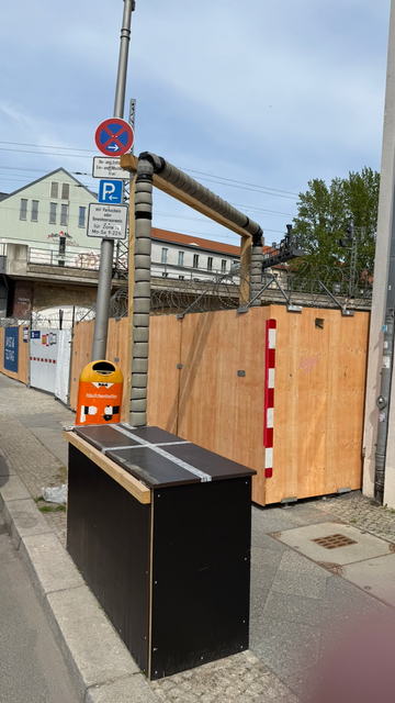 An empty lot at Luisenstr. in Berlin 'protected' by wooden panels with rolls of barbed wire on top. In front a wooden box which is easy to climb. Out of it comes a pipe covered in foam plastic supported by a wooden bridge that goes over the barbed wire and comes down again inside the lot.