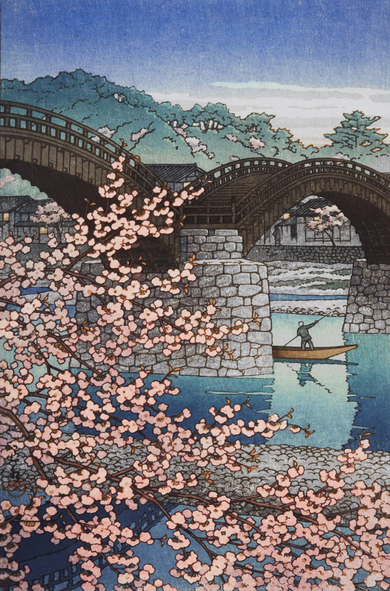 A lone oarsman pilots their small wooden boat along the calm Nishiki River, under the curving wooden arches of the Kintai Bridge. In the foreground, a cherry tree in bloom screens our view of the near side of the stony riverbank; beyond, houses backed by tall, healthy-looking green trees line up along the opposite side of the river. Above, a few wispy white clouds linger in a bright blue sky.