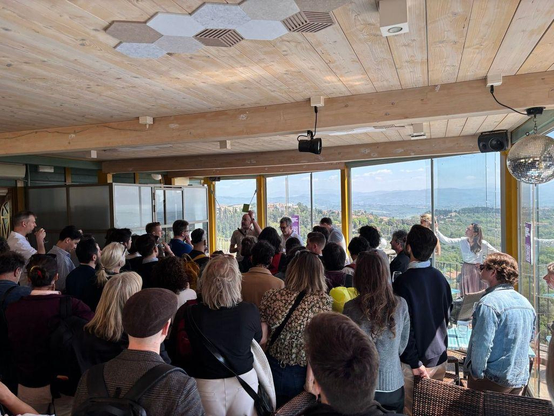 An image of a crowded room with rolling Italian hills in the background. 

Speaking to the room is Björn Staschen and Michael Foster. Björn is holding a small flyer up for the room to see. 