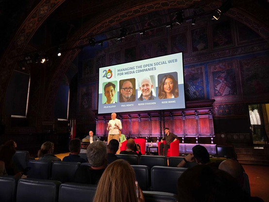 Björn Staschen speaks to the room in a Medieval hall. The walls are painted with various Italian family crests. He is joined on stage by Paul Nemitz, Rose Wang, and Felix Hlatky as part of a panel titled 'Managing the Open Social Web for Media Companies' at the International Journalism Festival.