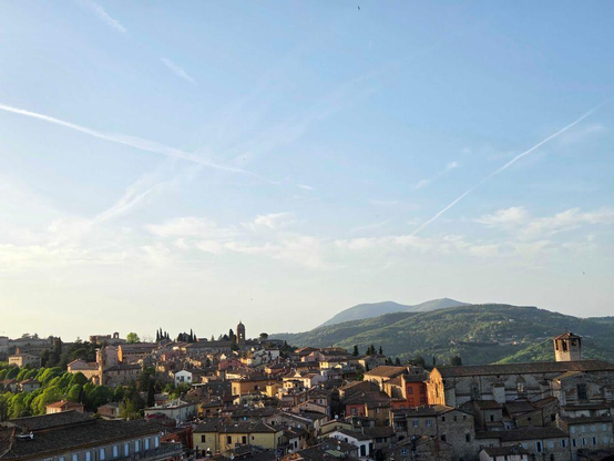 A landscape view of Perugia at sunset. In the foreground, old Italian buildings catch the light. In the background, tree covered hills rise up into the sky. 