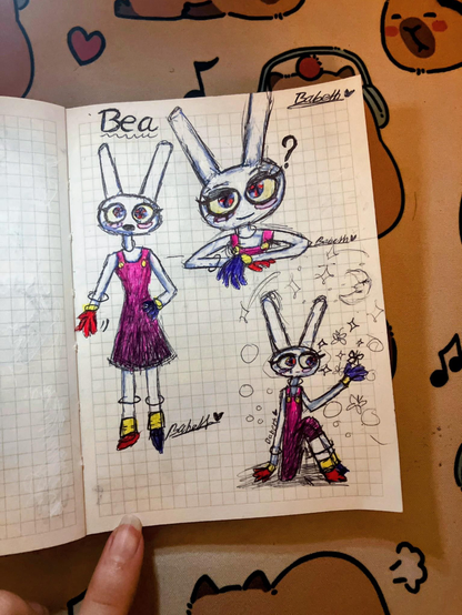 Photographie d'un carnet de croquis ouvert posé sur un sous-main décoré de visages d'animaux et de notes de musique. La page quadrillée présente trois dessins au stylo et aux crayons de couleur d'un lapin anthropomorphe blanc. Le prénom "Bea" est inscrit en haut à gauche.
 * Le dessin de gauche montre le personnage en pied, portant une robe violette à bretelles roses avec des boutons jaunes, ainsi que des gants et des chaussures aux couleurs asymétriques (rouge, jaune et bleu foncé).
 * Le dessin en haut à droite est un portrait à mi-corps du lapin avec une expression curieuse et un point d'interrogation à côté de sa tête.
 * Le dessin en bas à droite montre le lapin agenouillé, tendant la main vers une petite forme d'étoile ou de fleur, entouré de bulles dessinées, d'étincelles et d'un petit croissant de lune.
La signature "Babeth" accompagnée d'un cœur est visible à trois reprises sur la page. Le doigt d'une personne maintient le coin inférieur gauche du carnet.
