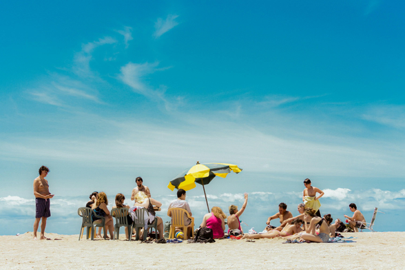 A group of people sitting and reclining on a sandy beach under a large yellow-and-green umbrella, with chairs arranged in a semicircle and a bright blue sky overhead. Photo by Emilce Giardino on Unsplash