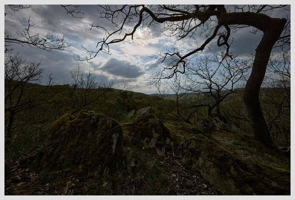 Wide angle landscape image. In the foreground we see a formation of three large rocks embedded in the ground, partly covered with moss. To the right, a leafless tree rises with its branches extending towards the center of the frame. Both rocks and tree are dark, almost silhouettes against the sky. The sky is a pale blue, with dark clouds moving in from the left, covering the sun in the center. Further in the background we see a forested range of hills.