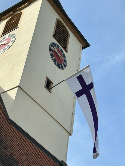 Ein lila-weiße Kreuzfahne weht an einem Kirchturm.