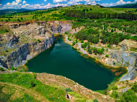 An aerial, high-angle view of a deep, emerald-green lake nestled inside a steep, rocky quarry. One side of the quarry features high, sheer cliffs of exposed gray and beige stone, while the other slopes gently upward, covered in vibrant green grass and scattered pine trees. Rolling green hills and a bright blue sky with fluffy white clouds stretch across the background.