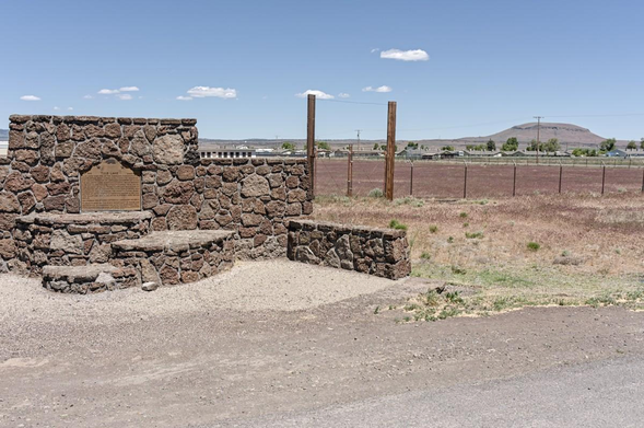 A color landscape photo of a monument made of large brown colored stones on the left. It has places to sit in front and a metal plaque on a wall in the back. To the right is a gate bordered by tall wood posts and a fence dividing the short weeds in front and a reddish gravel surface behind the fence. There are a few trees in the distance and a dome shaped hill in the background on the right. The sky is mostly clear, blue, with six small clouds.