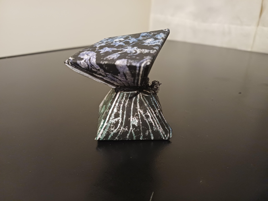 An hourglass-shaped sculpture formed from a lino print. It is pinched around the middle by a piece of black string. It shows flowers, either within or on it, from roots to blossoms.