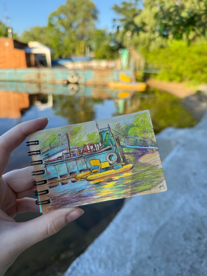 A hand holds up a small sketchbook with a colored pencil drawing of the real blue and yellow watercraft blurred in the background
