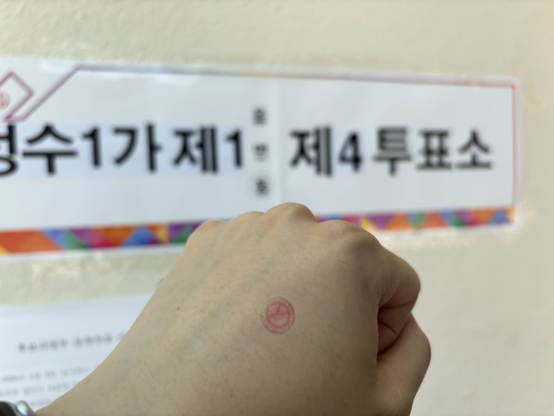 第9回 全國同時地方選擧 投票 認證 寫眞. 背景에는 「聖水1街第1洞 第4投票所」라고 적힌 案內板이 附着되어 있고, 그 앞쪽으로 붉은색 投票 圖章(卜 模樣)이 鮮明하게 찍힌 揭示者 洪民憙의 오른손 주먹이 놓여 있다.