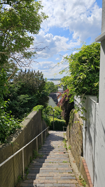 Schmale Treppe mit Pflastersteinen zwischen einer Mauer links und einer Steinmauer mit weißem Zaun rechts. Beidseitig dichter Bewuchs mit Bäumen, Sträuchern und Kletterpflanzen. Im Hintergrund sind Dächer von Häusern sowie ein Gewässer sichtbar. Darüber blauer Himmel mit großen Wolken.