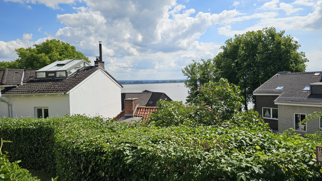 Blick über eine dichte Hecke auf mehrere Häuser mit geneigten Dächern. Zwischen den Gebäuden ist ein breites Gewässer sichtbar. Im Vordergrund und Hintergrund stehen Bäume und Sträucher. Darüber blauer Himmel mit großen Wolken.