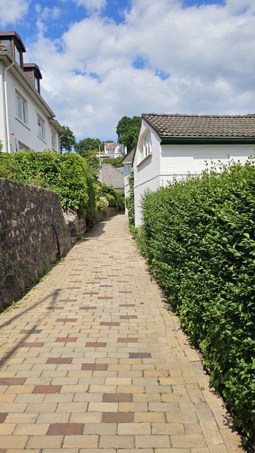 Gepflasterter Weg zwischen einer Steinmauer mit Hecke auf der linken Seite und einem weißen Gebäude mit Hecke auf der rechten Seite. Der Weg verläuft leicht ansteigend und führt zwischen weiteren Gebäuden hindurch. Im Hintergrund sind Häuser, Bäume und Sträucher zu sehen. Darüber ein teilweise bewölkter Himmel.