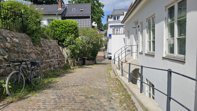 Kopfsteingepflasterter Weg zwischen einer Natursteinmauer mit Zaun auf der linken Seite und einem weißen Gebäude auf der rechten Seite. Am linken Rand steht ein Fahrrad. Entlang des Weges befinden sich Hecken, Sträucher und weitere Gebäude. Im Hintergrund verläuft der Weg zwischen Häusern weiter. Darüber sind Bäume und ein teilweise bewölkter Himmel zu sehen.