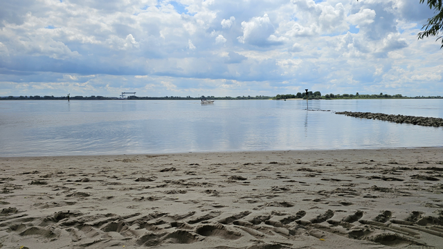 Sandstrand am Ufer eines breiten Gewässers. Im Vordergrund sind Fußspuren und Unebenheiten im Sand zu sehen. Auf dem Wasser fährt ein kleines Boot. Rechts ragt eine niedrige Steinaufschüttung mit Schifffahrtszeichen ins Wasser. Am gegenüberliegenden Ufer sind Bäume und flache Vegetation sichtbar. Darüber ein Himmel mit zahlreichen Wolken und einzelnen blauen Bereichen.