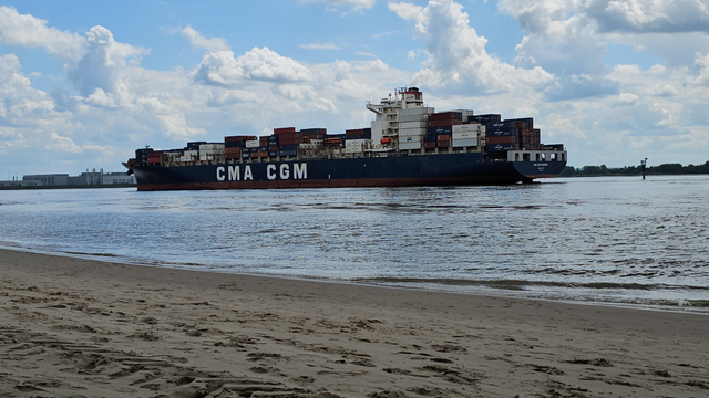 Sandstrand am Ufer eines breiten Gewässers. Auf dem Wasser fährt ein großes Containerschiff mit gestapelten Containern. Im Vordergrund sind Fußspuren im Sand zu sehen. Am gegenüberliegenden Ufer befinden sich Gebäude und flache Vegetation. Darüber ein Himmel mit zahlreichen Wolken und einzelnen blauen Bereichen.