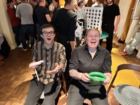 Tommi, a young white man, reacts with an expression of surprise whilst holding a Wii controlled. Beside him, Saskia, a young white woman, is holding a green steering wheel Wii controller and shouting. 