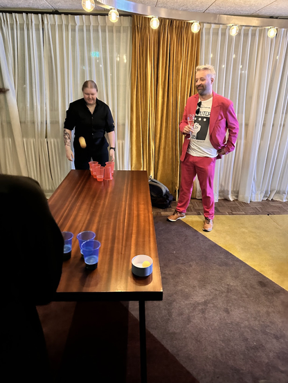 Beer pong is being played on a long table. A yellow ping pong ball is midair with Saskia, a young white woman dressed in all black, and Robin, a middle-aged white man in a hot pink suit, watching it. 