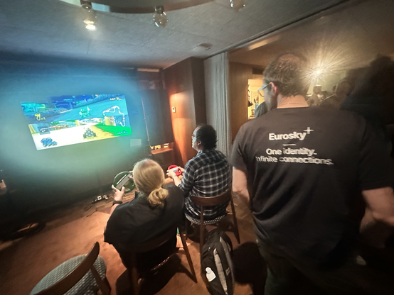 A screen can be seen with Mario Kart being played. One player is in first position, the other in twelth. The players are Saskia, a young white woman dressed in black, and Ben, a middle-aged white man with black hair and wearing a check shirt. The photo is taken from behind them so their expressions can't be seen. In the foreground, a white man can be seen wearing a Eurosky t-shirt, reading 'Eurosky - One identity. Infinite connections.' 