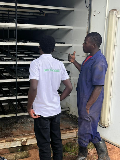 A farm worker explains a hydroponic fodder growing chamber with multiple trays, while a visitor listens and observes the system during a farm tour.