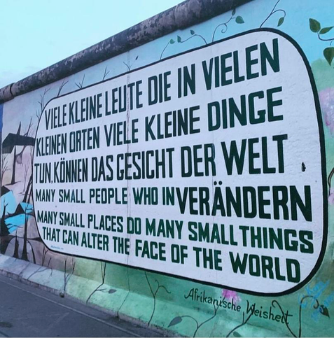 A mural on the Berlin Wall featuring a quote in German and English, emphasizing the impact of small people and places on the world. The text is bold and prominently displayed, with a backdrop of a painted wall.
